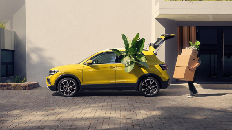 Un VW T-Cross jaune est garée, remplie de plantes. Un homme en plein déménagement remplit le coffre de cartons