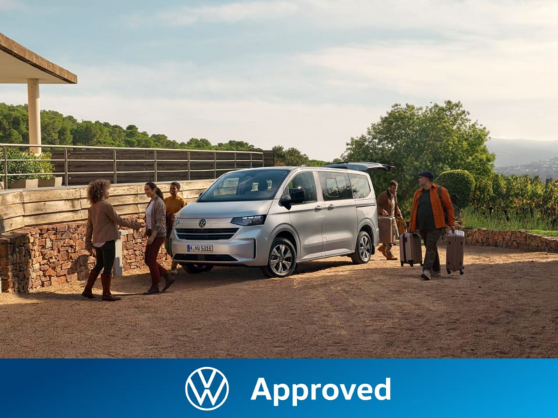 Personas con maletas alrededor de una furgoneta Approved de Volkswagen aparcada en una casa de campo