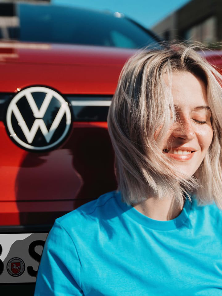 Una chica sentada delante de un Volkswagen con el logo del coche de fondo