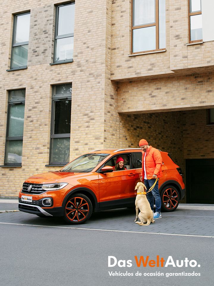 Una pareja con un perro y un Volkswagen T-Cross naranja frente a un edificio de ladrillo