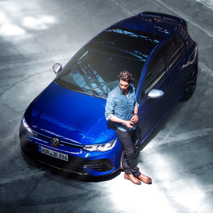 Chico apoyado en el capó de un Volkswagen Golf R azul visto desde arriba
