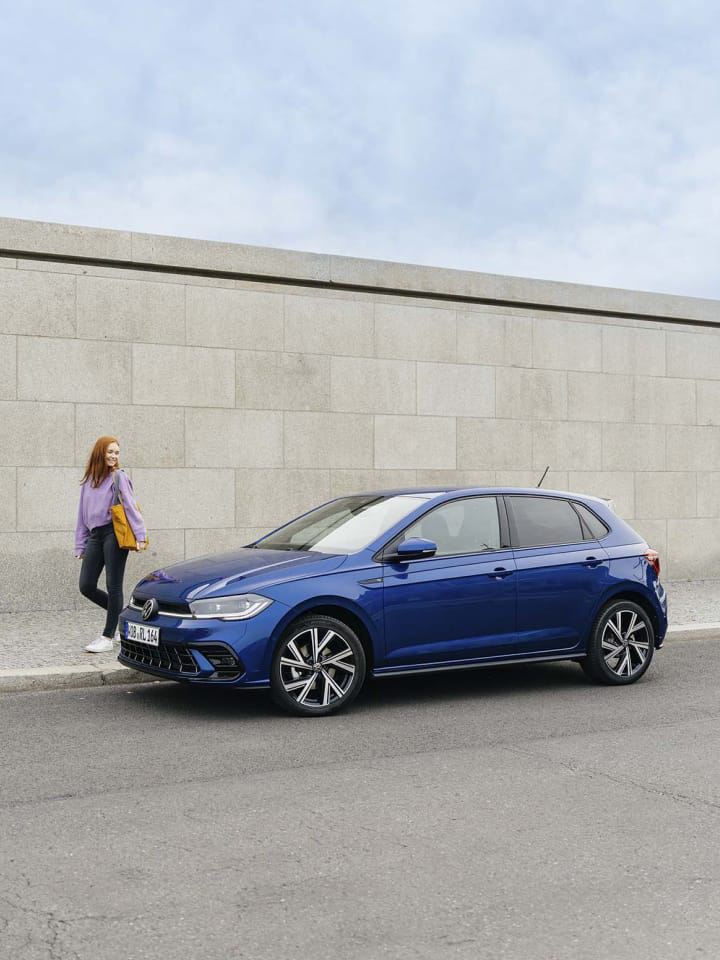 Una chica junto a un Volkswagen Polo azul aparcado frente a un muro de concreto