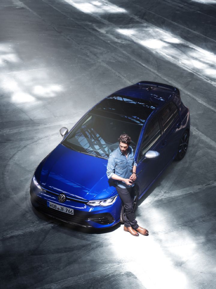 Chico apoyado en el capó de un Volkswagen Golf R azul visto desde arriba