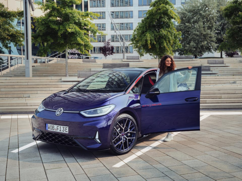 Una persona saliendo de su coche eléctrico de Volkswagen que está aparcado en un estacionamiento al aire libre, al fondo árboles y edificios