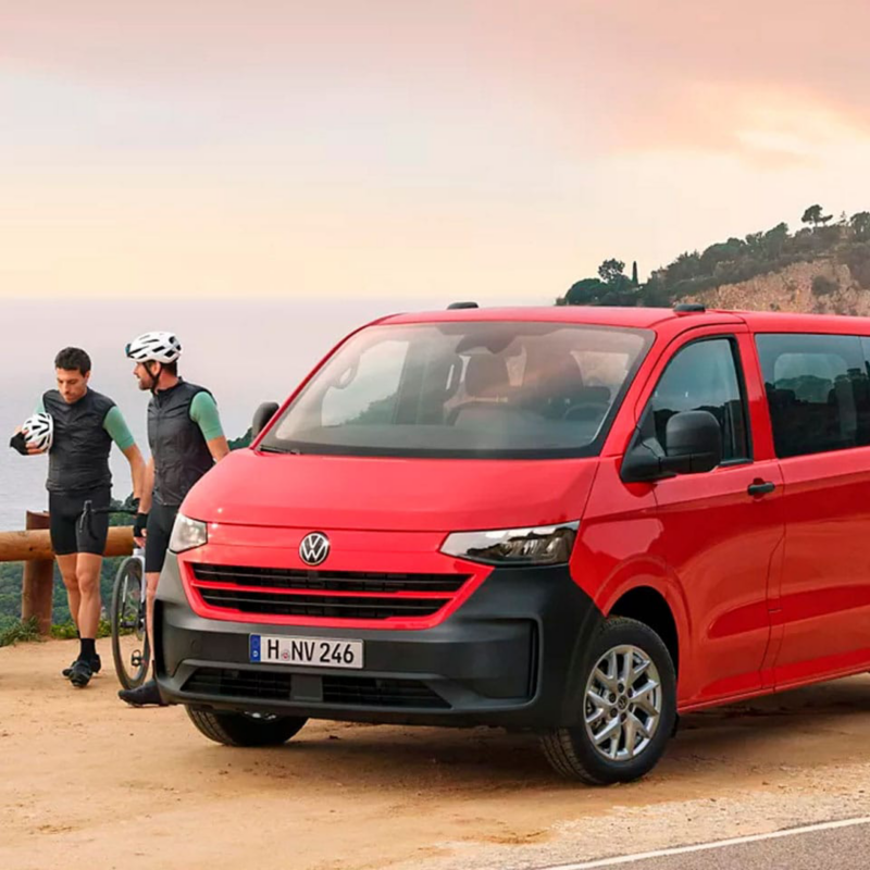 Una furgo roja de Volkswagen aparcada en un atardecer con vistas al mar y dos ciclistas caminando al lado