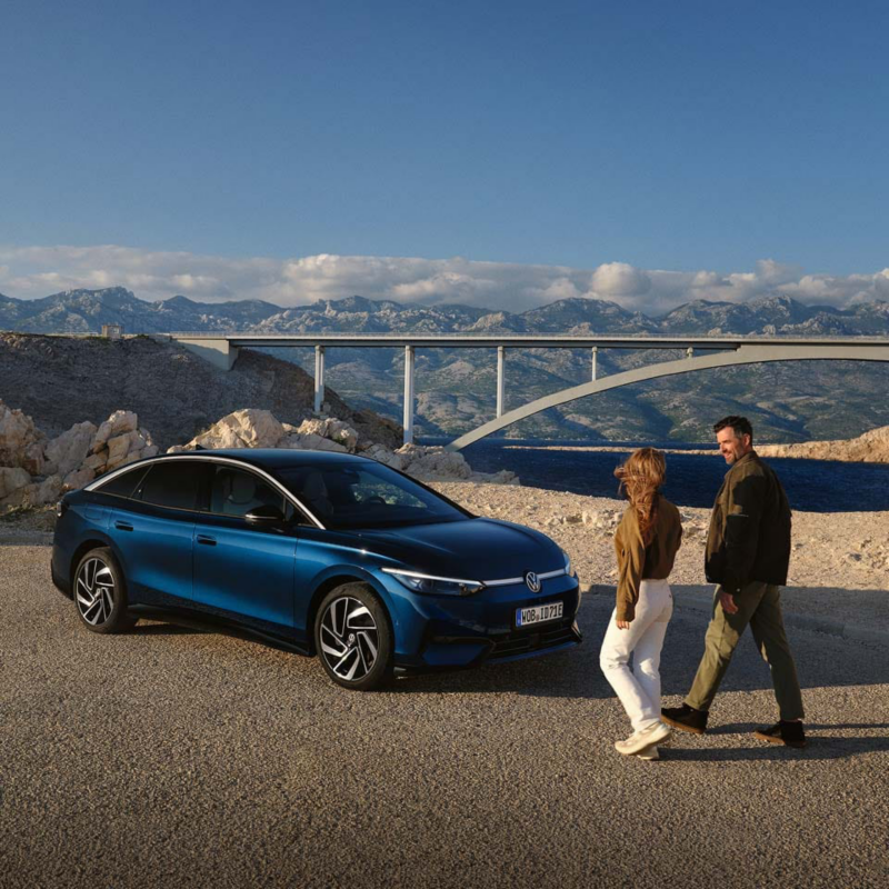 Dos personas junto a un Volkswagen ID.7 azul en un paisaje con un puente