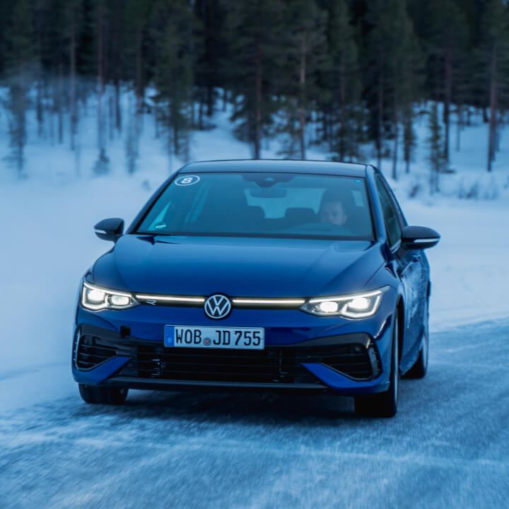 Un Volkswagen Polo azul en una carretera nevada con árboles de fondo. Tiene las luces delanteras encendidas.