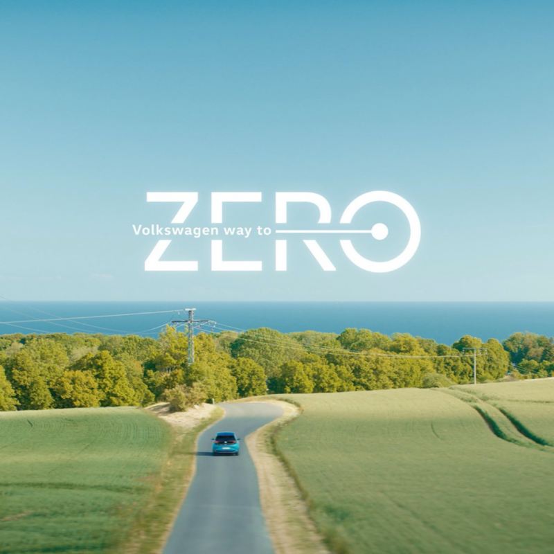 Un Volkswagen visto de frente circulando por una carretera en el campo y el mar de fondo con el logo de Way to Zero sobre impreso