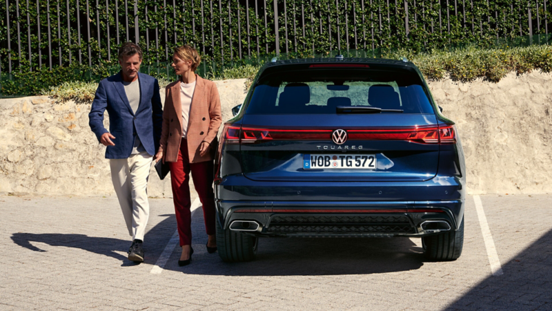 Ein Mann und eine Frau laufen an einem blauen VW Touareg R eHybrid vorbei.