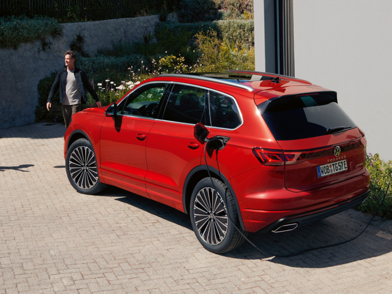 Ein roter VW Touareg Elegance eHybrid wird geladen. Ein Mann läuft vor dem Wagen.