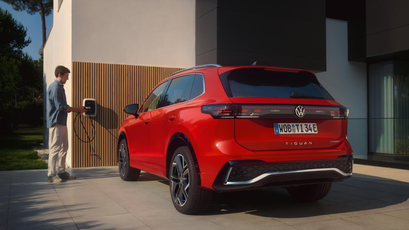 A red Volkswagen Tiguan SUV is parked in front of a wall with a man using a Ohme wallbox to charge it.