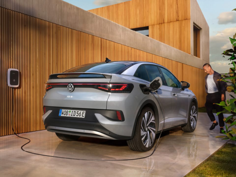 Un hombre junto a un Volkswagen de color gris conectado a carga estacionado en el exterior de un casa