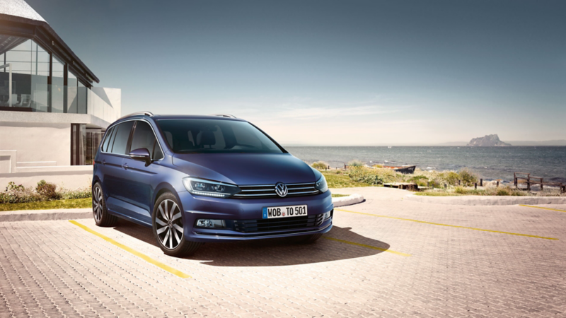 Ein blauer VW Touran steht auf einem Parkplatz, im Hintergrund Meer und Strand.