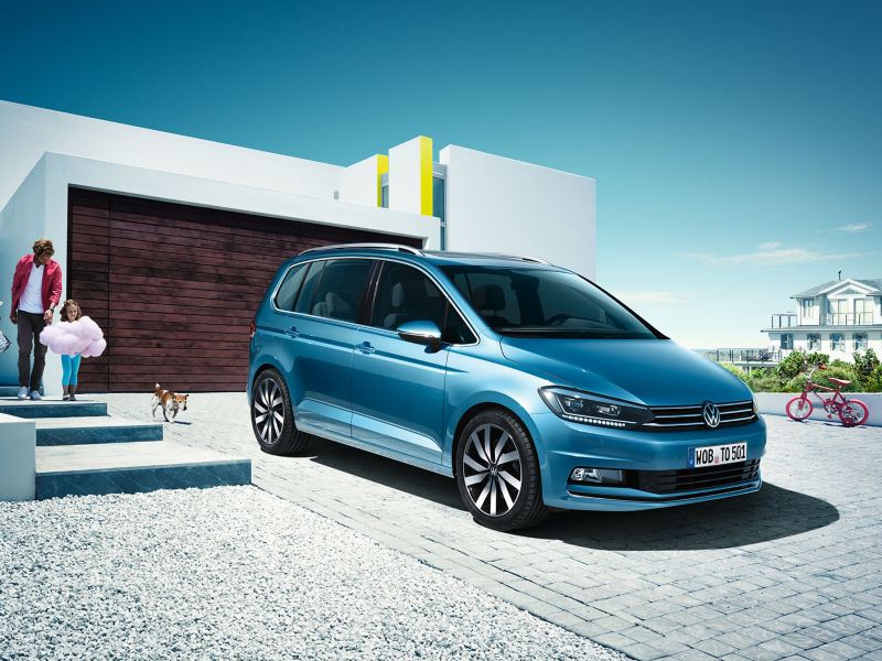 Sininen Volkswagen Touran talon edustalla pysäköitynä ja kuvattuna etuviistosta. Auton takana on perhe kävelemässä