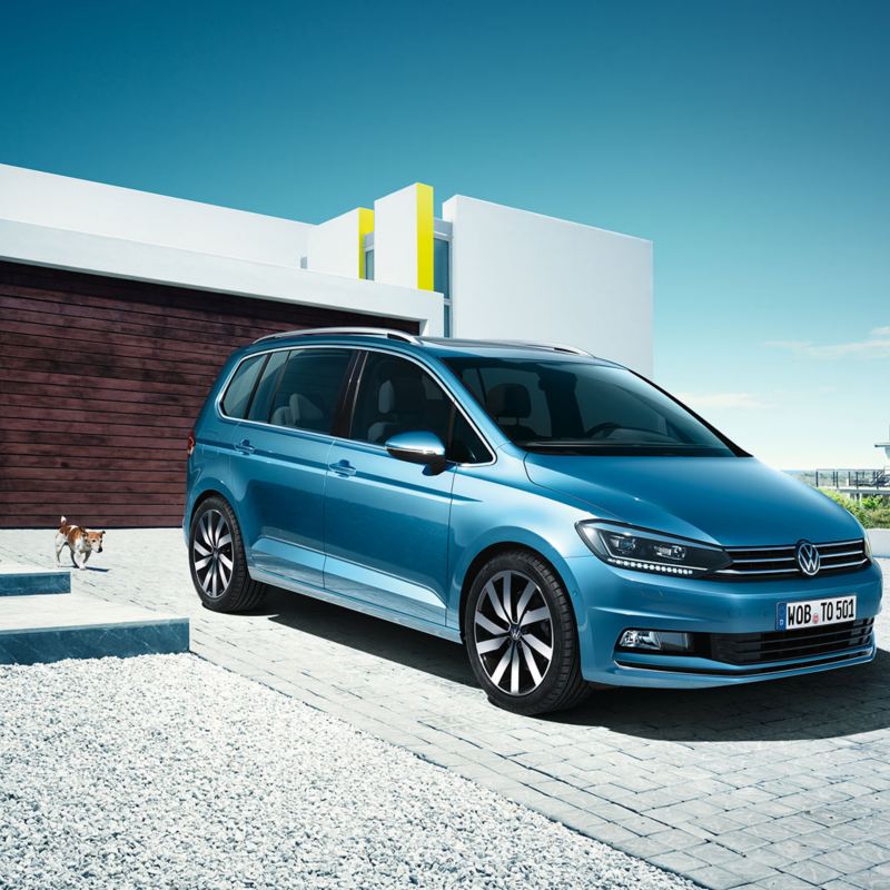 Sininen Volkswagen Touran talon edustalla pysäköitynä ja kuvattuna etuviistosta. Auton takana on perhe kävelemässä