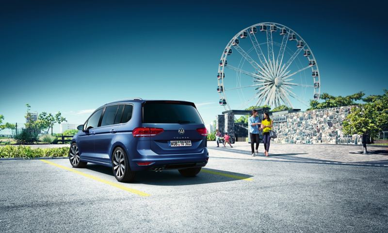 Une famille quitte un parc d'attraction avec une grande roue, un VW Touran bleu garé et vu de l'arrière est au premier plan.