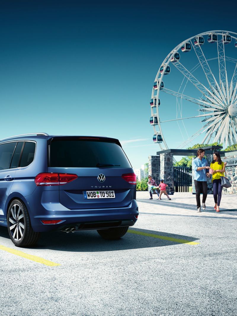 Une famille quitte un parc d'attraction avec une grande roue, un VW Touran bleu garé et vu de l'arrière est au premier plan.