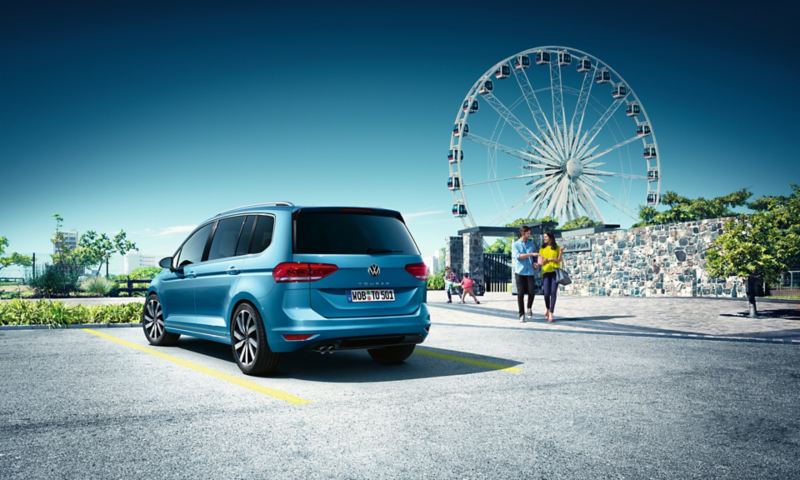 Familie verlässt einen Vergnügungspark mit Riesenrad, im Vordergrund steht ein blauer VW Touran.