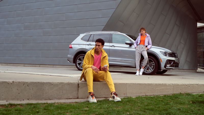 Une jeune femme est appuyée sur un Tiguan 2022 de Volkswagen argent pyrite métallisé, tandis qu’un homme est assis sur le trottoir avec un téléphone intelligent.