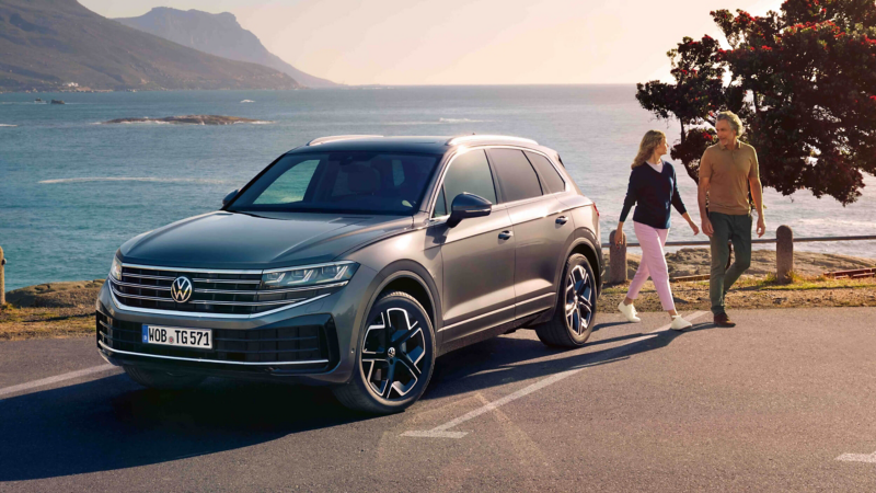A man and woman walking behind a parked VW Touareg in a seaside setting