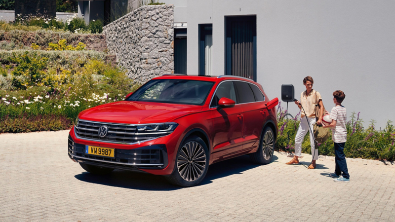 Un Touareg rouge est garé devant la maison, et une mère s'apprête à le charger à la borne de recharge
