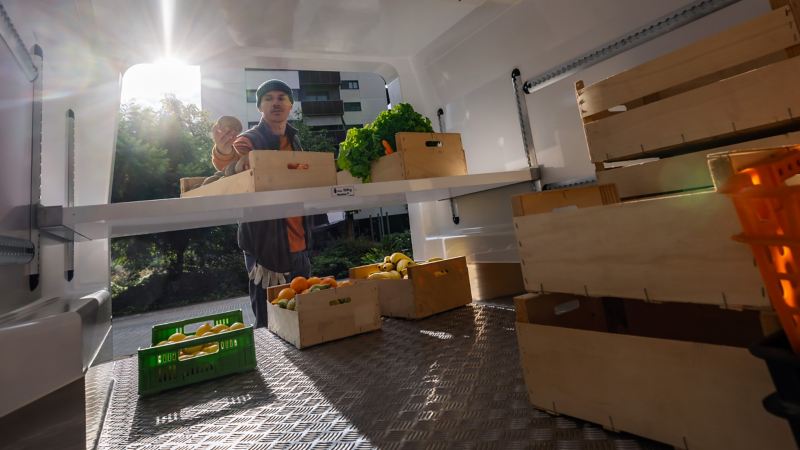 Ein Lieferant steht an der geöffneten Heckklappe eines VW Transporters und begutachtet geladene Obstkisten.