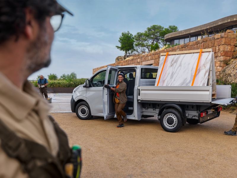 VW Transporter Pritschenwagen beladen mit zwei Marmorplatten vor einem modernen Gebäude in mediterraner Landschaft. Mehrere Handwerker befinden sich um das Fahrzeug.