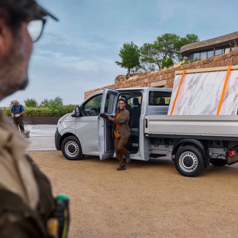 VW Transporter Pritschenwagen beladen mit zwei Marmorplatten vor einem modernen Gebäude in mediterraner Landschaft. Mehrere Handwerker befinden sich um das Fahrzeug.