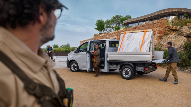 VW Transporter Pritschenwagen beladen mit zwei Marmorplatten vor einem modernen Gebäude in mediterraner Landschaft. Mehrere Handwerker befinden sich um das Fahrzeug.