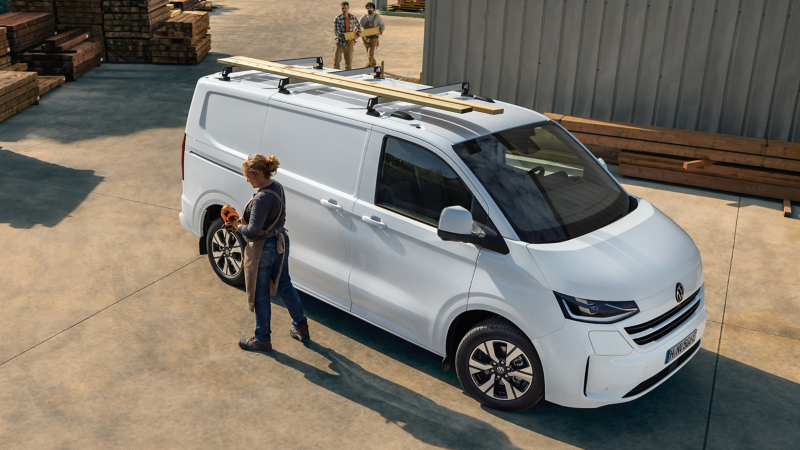 Eine Bauarbeiterin steht vor einem weißen VW Kastenwagen der auf dem Dach Holzbalken transportiert.