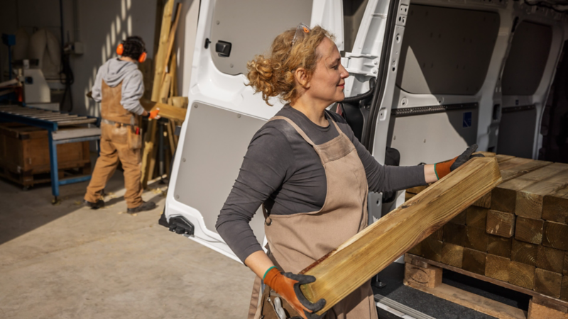 Eine Handwerkerin verstaut Holzpfosten auf der Ladefläche des VW Transporter Kastenwagen.
