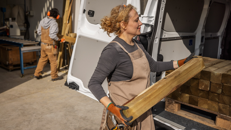 Eine Handwerkerin verstaut Holzpfosten auf der Ladefläche des VW Transporter Kastenwagen.