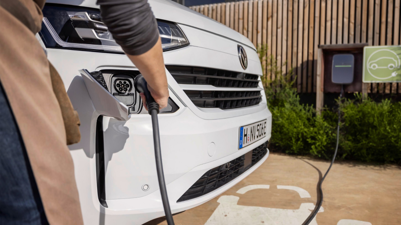 Eine Person steckt das Ladekabel in den Frontanschluss eines weißen Elektro-Transporters von Volkswagen an einer Ladestation im Freien.