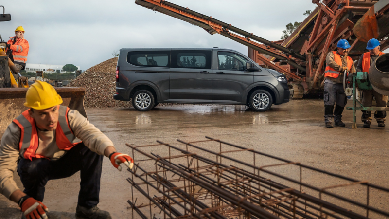 Ein dunkelgrauer VW Transporter Kombi steht auf einer Baustelle, während im Vordergrund und Hintergrund Arbeiter mit Bauarbeiten beschäftigt sind.