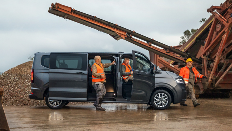 Bauarbeiter steigen aus einem VW e-Transporter Kombi aus. Im Hintergrund stehen große Maschinen.