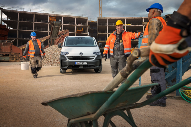 Ein VW Transporter steht auf einer Baustelle, im Vordergrund laufen Bauarbeiter herum