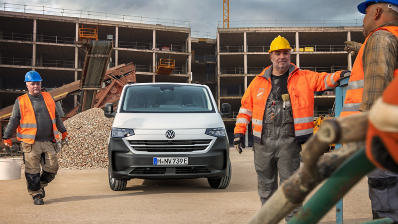 VW e-Transporter Kombi umgeben von Bauarbeitern auf einer Baustelle.