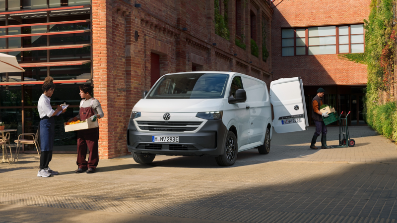 VW e-Transporter parkt neben einem Café. Lieferanten entladen Obst- und Gemüsekisten aus dem Heck des Fahrzeugs. Die Besitzerin des Cafés quittiert den Empfang der Ware.