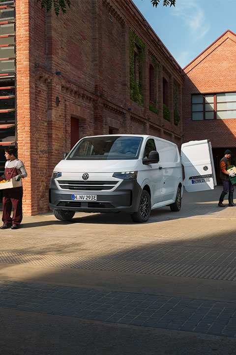  Un fourgon Volkswagen Transporter blanc est garé dans une rue pavée, devant des bâtiments en briques rouges. L'homme sur le côté gauche de l'image porte une boîte tandis que l'homme sur le côté droit de l'image est près de la porte arrière du véhicule.