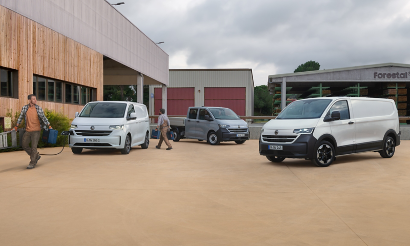 Drei verschiedene Varianten des neuen VW Transporters stehen auf dem Betriebsgelände einer Firma, während ein Mann ein Ladekabel herauszieht.