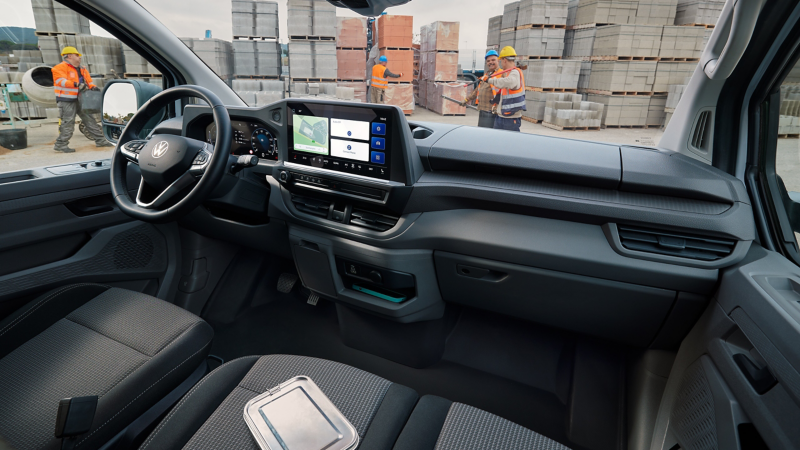 Die Innenansicht des VW Transporter Cockpits zeigt das moderne Infotainment-Display, während im Hintergrund lachende Bauarbeiter zu sehen sind.