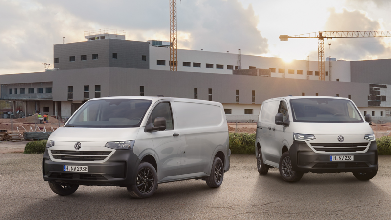 Zwei hellgraue Volkswagen Transporter Kastenwagen stehen vor einer Großbaustelle mit Kränen bei Sonnenuntergang.