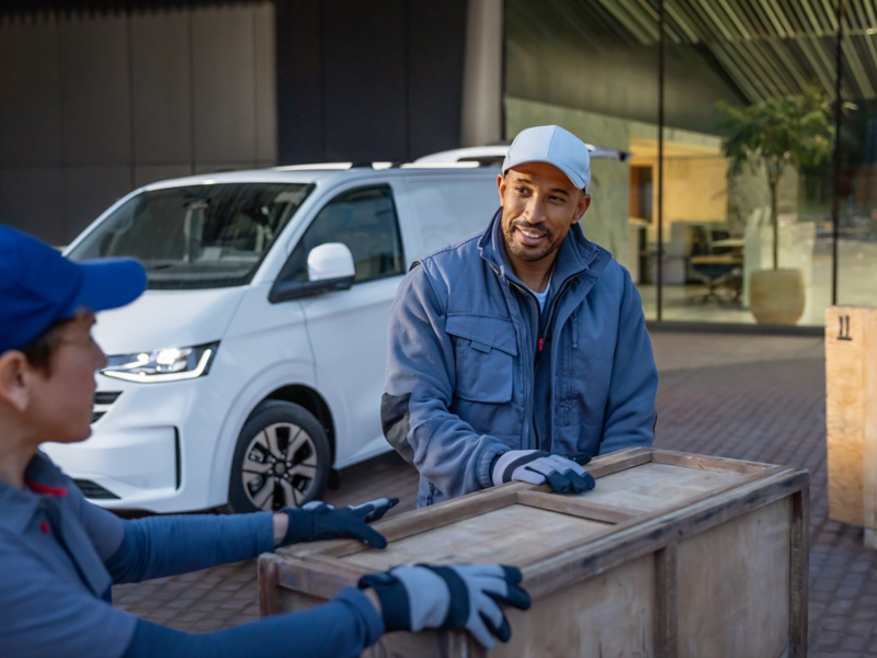 Zwei Kuriere liefern eine Holzkiste ab, im Hintergrund steht ein VW Transporter.