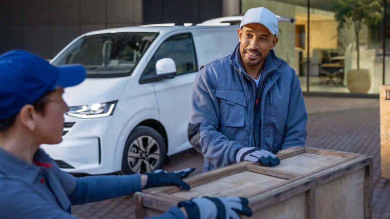 Zwei Kuriere stehen neben großen Holzkisten, im Hintergrund steht ein VW Transporter Kastenwagen mit geöffneter Heckklappe.