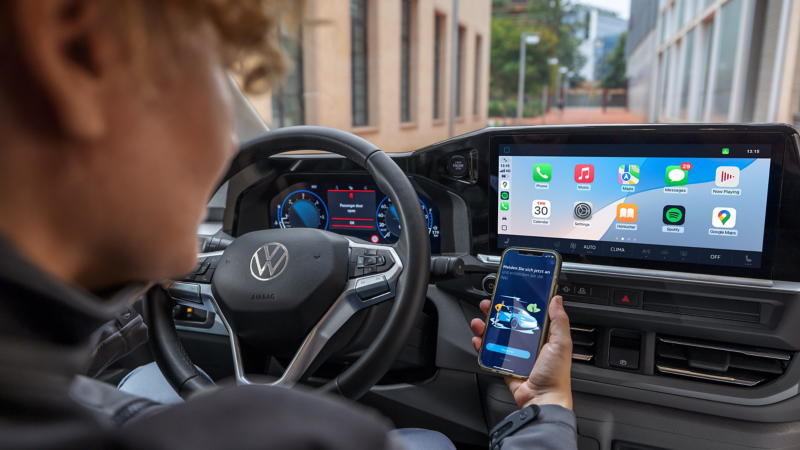 Eine Person am Steuer eines parkenden VW Transporter blickt auf ein Smartphone mit geöffneter VW Connect App.