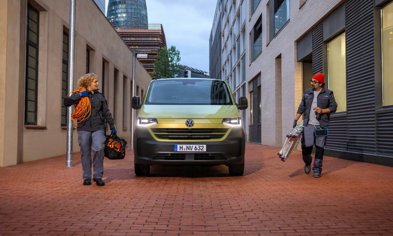 Ein VW Transporter parkt in einer Seitenstraße einer Stadt und zwei Handwerker mit Werkzeug laufen daneben her.