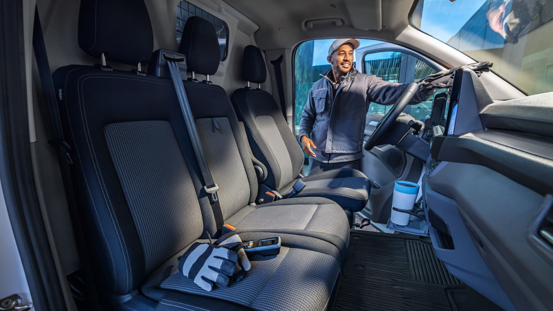 Ein Paketzusteller steht in der Tür eines VW Transporter, auf dem Beifahrersitz liegen Handscanner und Handschuhe.