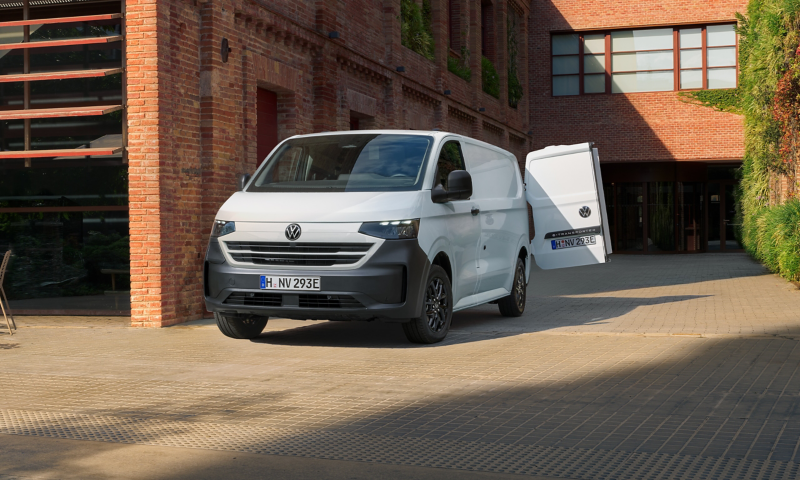 Ein VW Transporter mit offenen Heckklappen steht neben einer alten Lagerhalle in der Sonne.