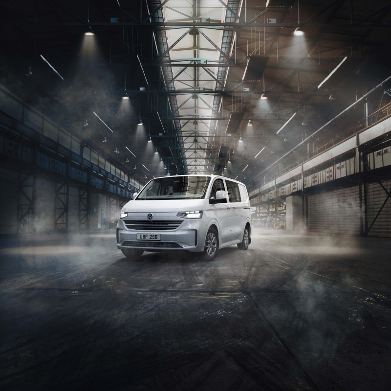 White Transporter Kombi van parked in a large, dimly lit industrial warehouse. Overhead lights cast dramatic shadows, creating a mysterious and futuristic atmosphere.