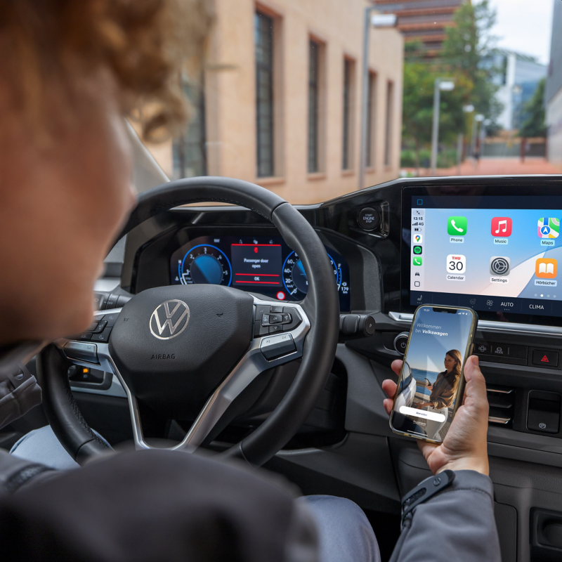 En person vid ratten i en parkerad VW Transporter tittar på en smartphone med VW Connect-appen öppen.